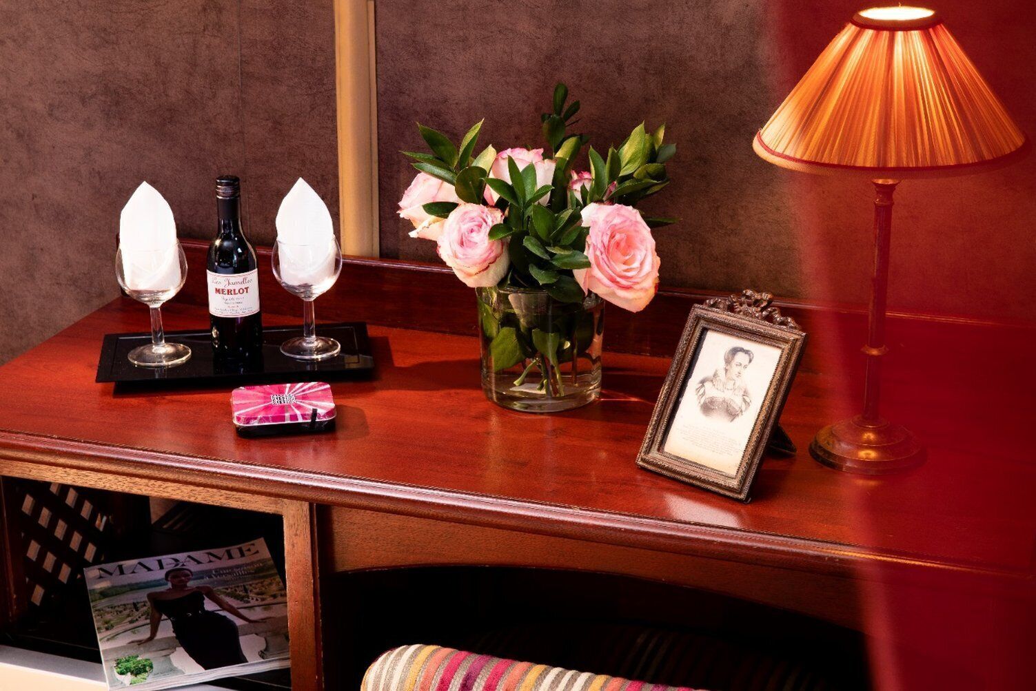 A wooden table with a wine bottle, two glasses, a vase with pink roses, a framed picture, and a lamp.