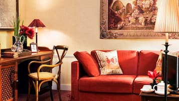 A cozy living room with a red sofa, a wooden chair, a small table with a lamp, and a framed painting on the wall.