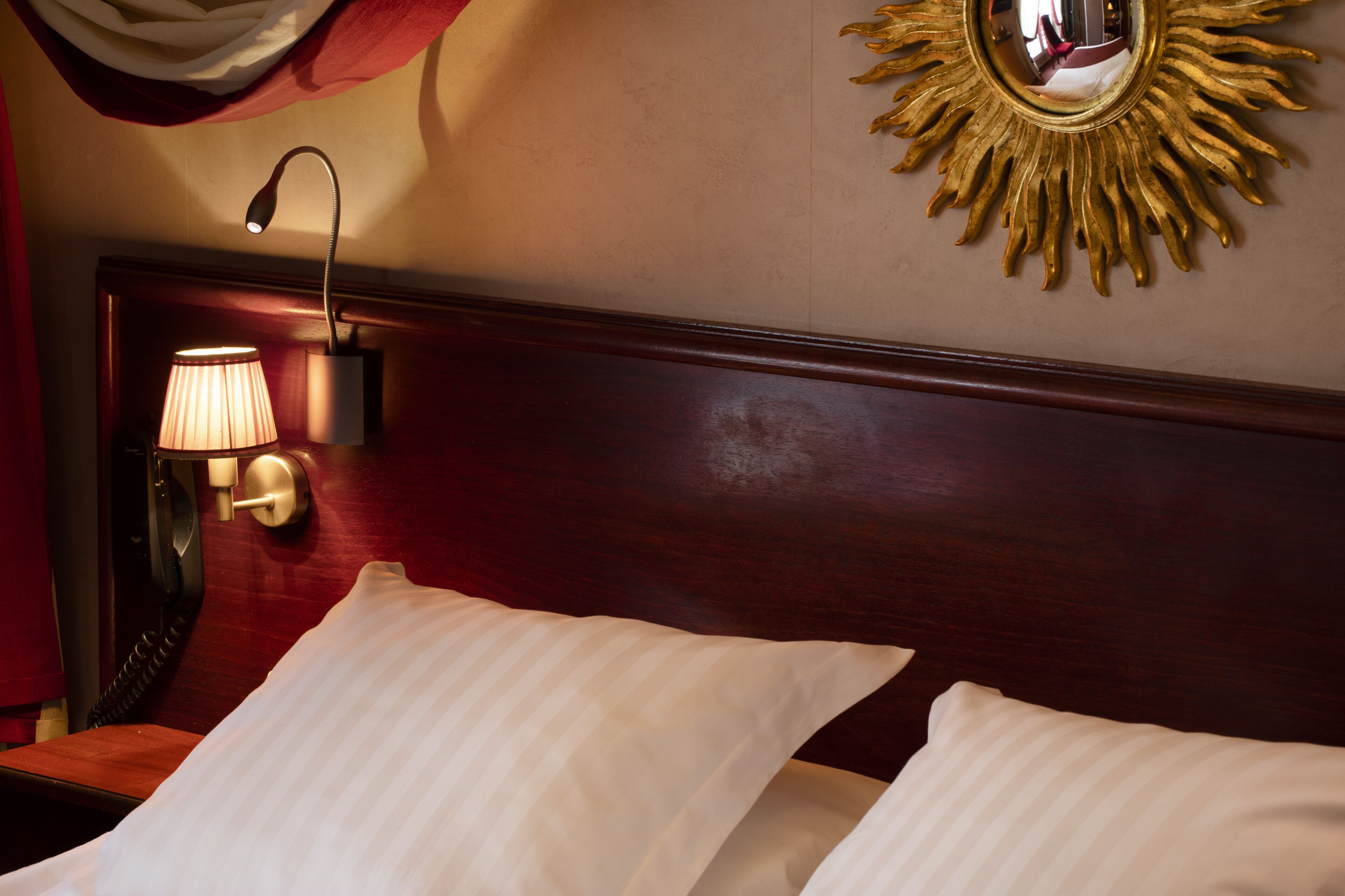 A hotel room with a wooden headboard, a wall-mounted lamp, and a sunburst mirror.
