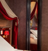A hotel room with a large mirror, red and white drapes, a bedside lamp, and a bed with white linens.