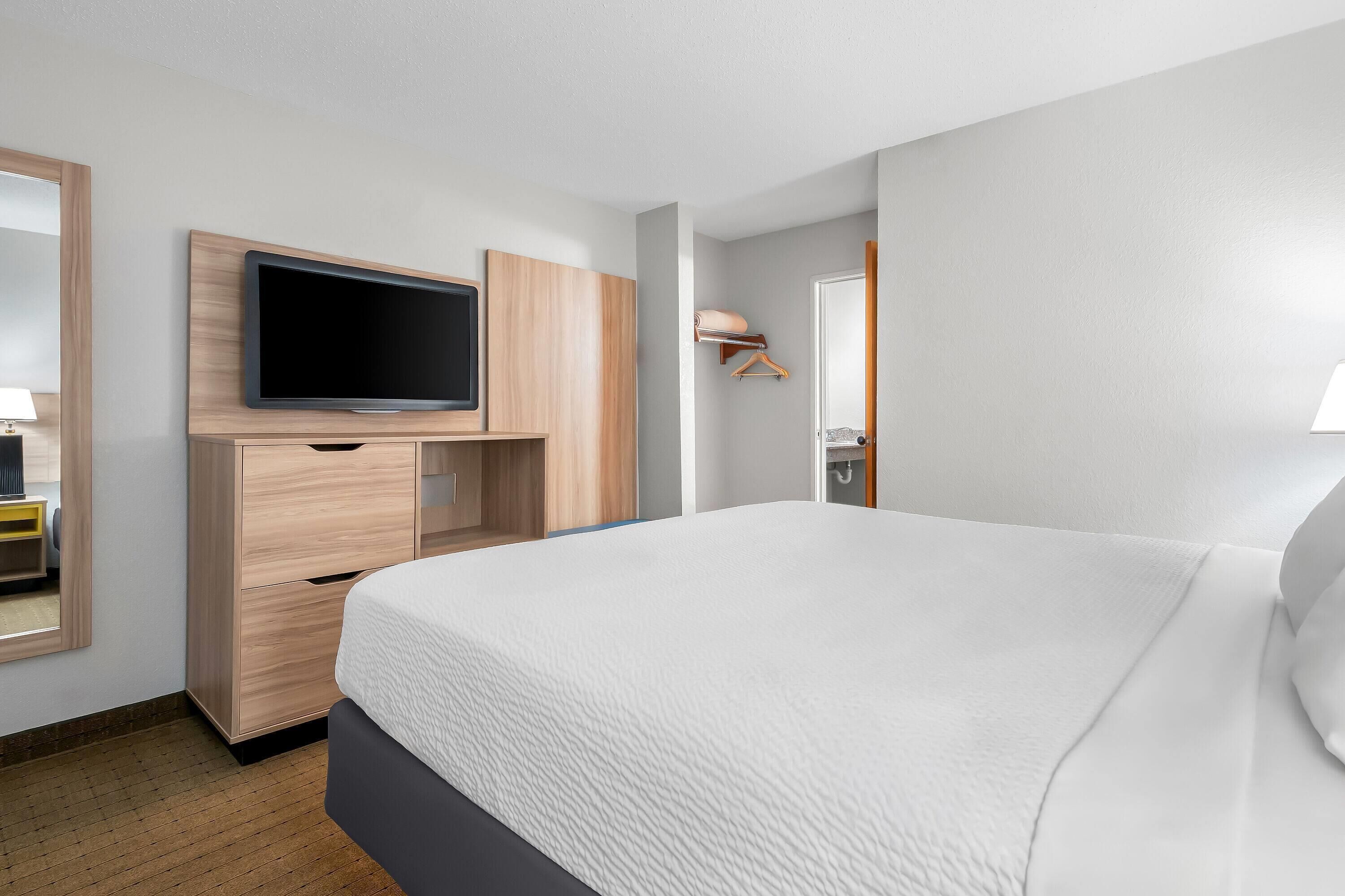 Una habitación de hotel moderna con una cama grande, un televisor de pantalla plana montado en un mueble de madera y una mesita de noche con una lámpara.