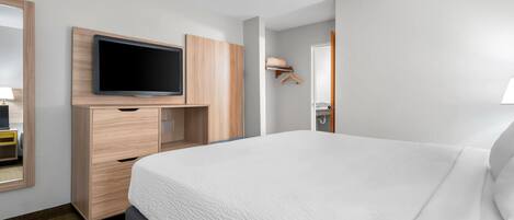 A modern hotel room with a large bed, a flat-screen TV mounted on a wooden cabinet, and a small bedside table with a lamp.