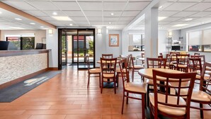 A hotel lobby with a reception desk, multiple dining tables and chairs, and a view of the outside through large windows.