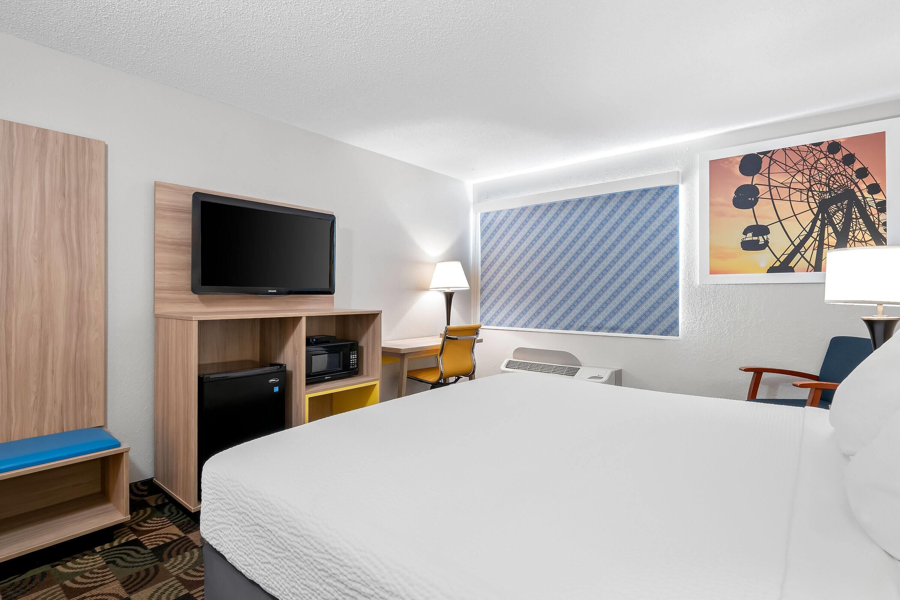 Habitación de hotel con cama, televisión, escritorio con lámpara, una silla y un cuadro de noria en la pared.