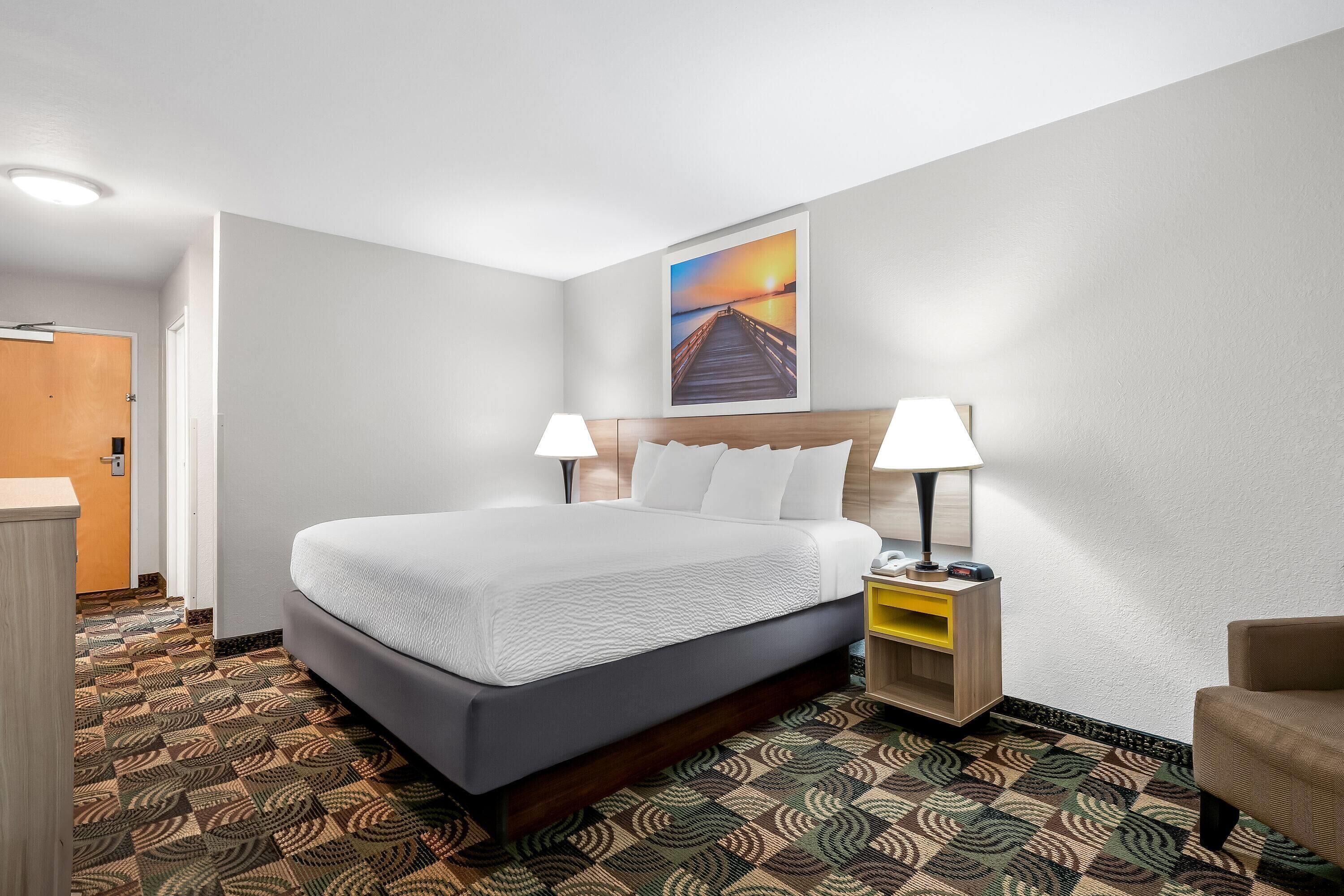 Una habitación de hotel con una cama grande, dos lámparas de noche, un mesita de noche, cabecera de madera y alfombra estampada.