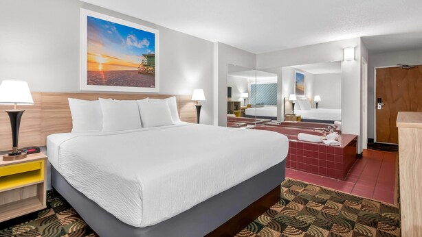 A modern hotel room with a large bed, a mirror, a bathroom with a red bathtub, and a painting of a sunset on the wall.