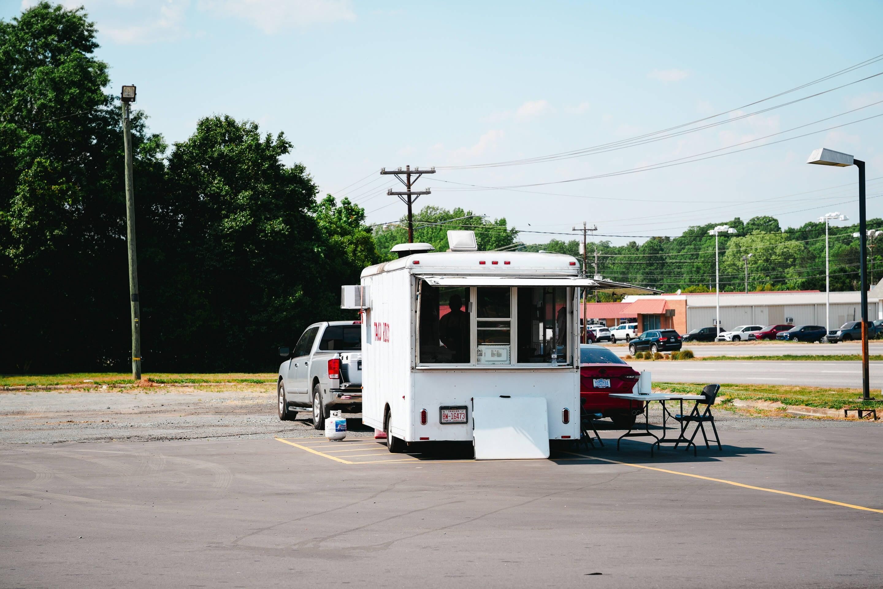 A food truck parked in a parking lot with a table and chairs, surrounded by trees and buildings.