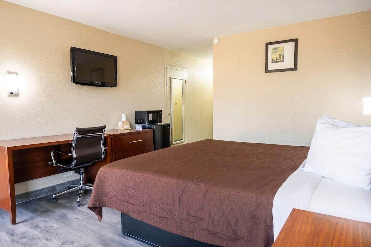 A hotel room with a bed, a desk with a chair, a television, a door, and a framed picture on the wall.