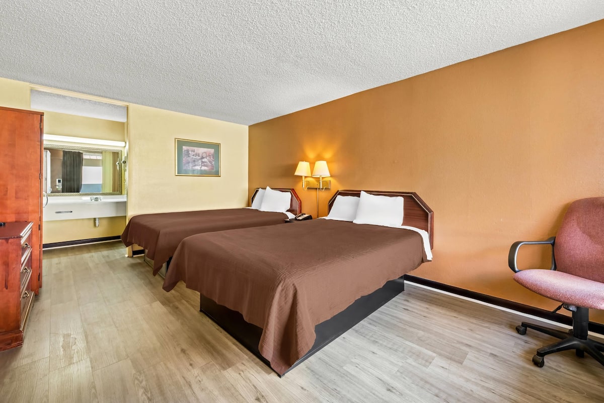A hotel room with two beds, a desk, a chair, a mirror, and a wall-mounted lamp.