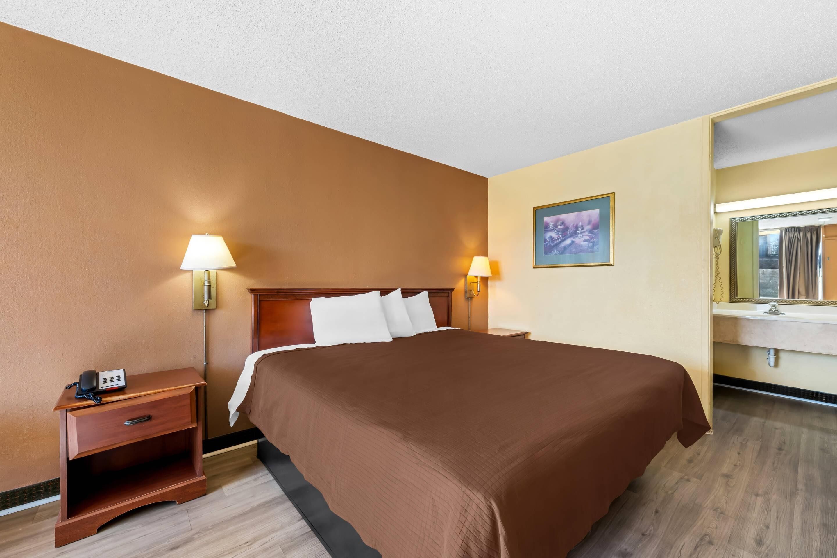 A hotel room with a large bed, bedside lamps, a nightstand with a phone, a painting on the wall, and a mirror above the sink.