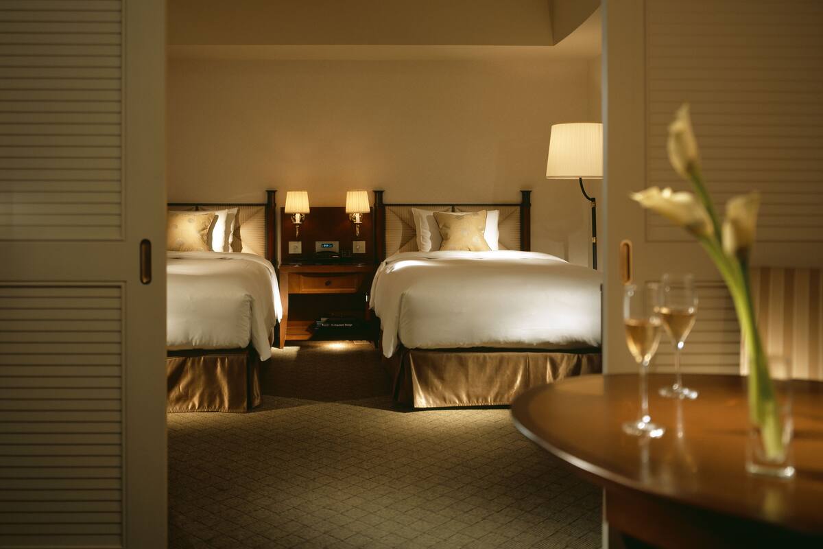 A hotel room with two beds, a nightstand with lamps, a small table with a glass of wine, and a door.