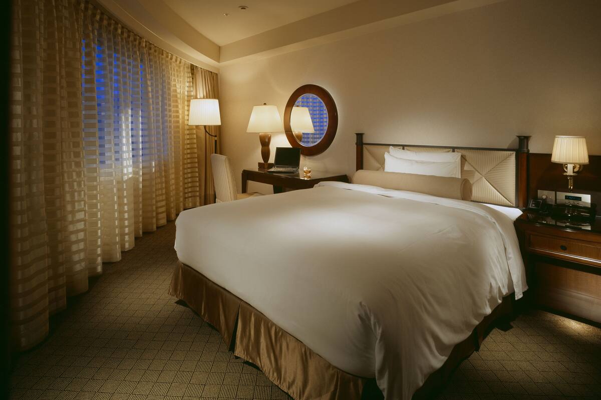 A hotel room with a large bed, two bedside tables, a desk, a chair, a lamp, and a window with curtains.