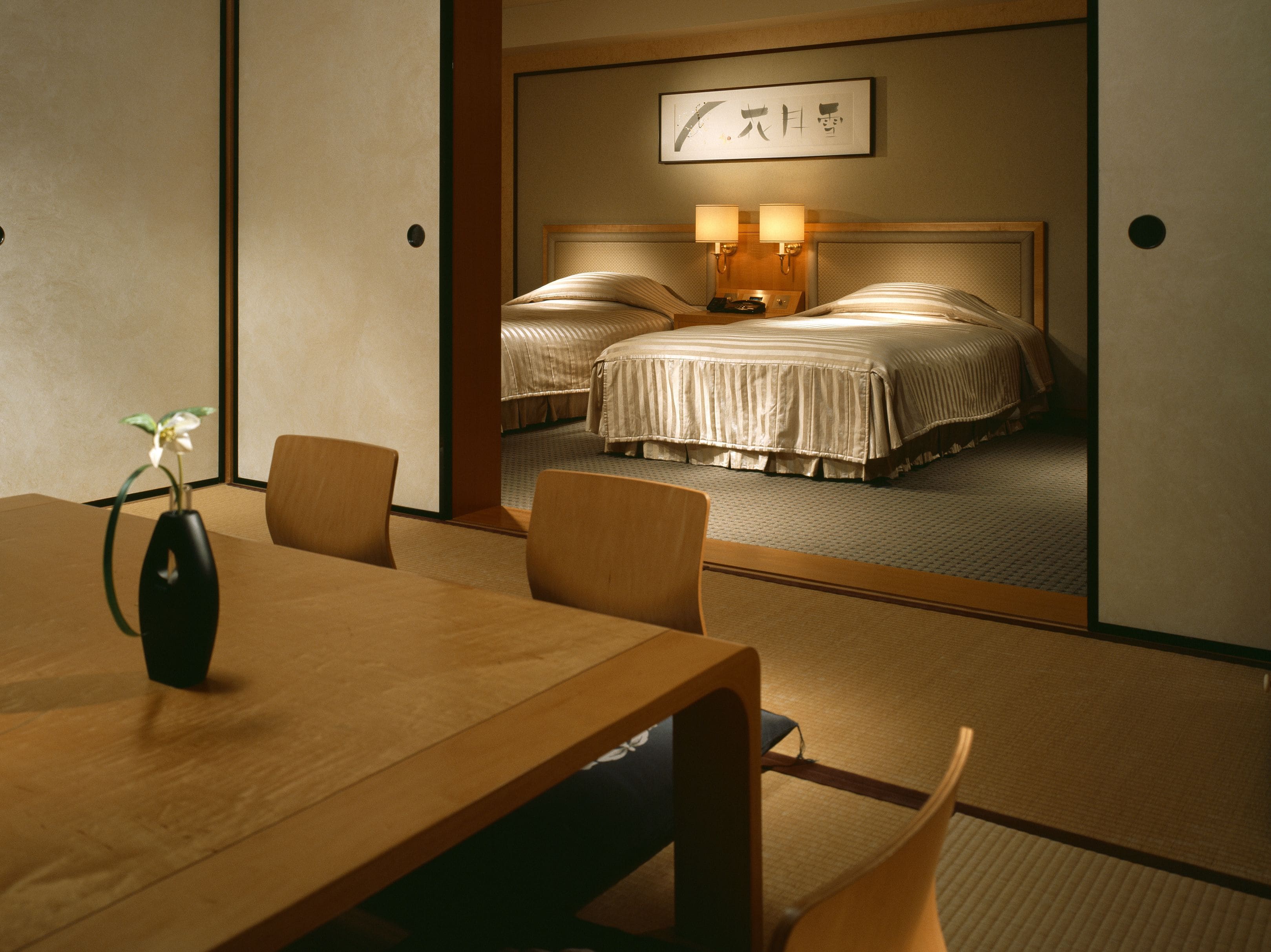Une chambre d’hôtel au décor japonais traditionnel, avec un sol recouvert de tatamis, une basse table en bois entourée de chaises et un lit doté d’une tête de lit et de lampes de chevet.