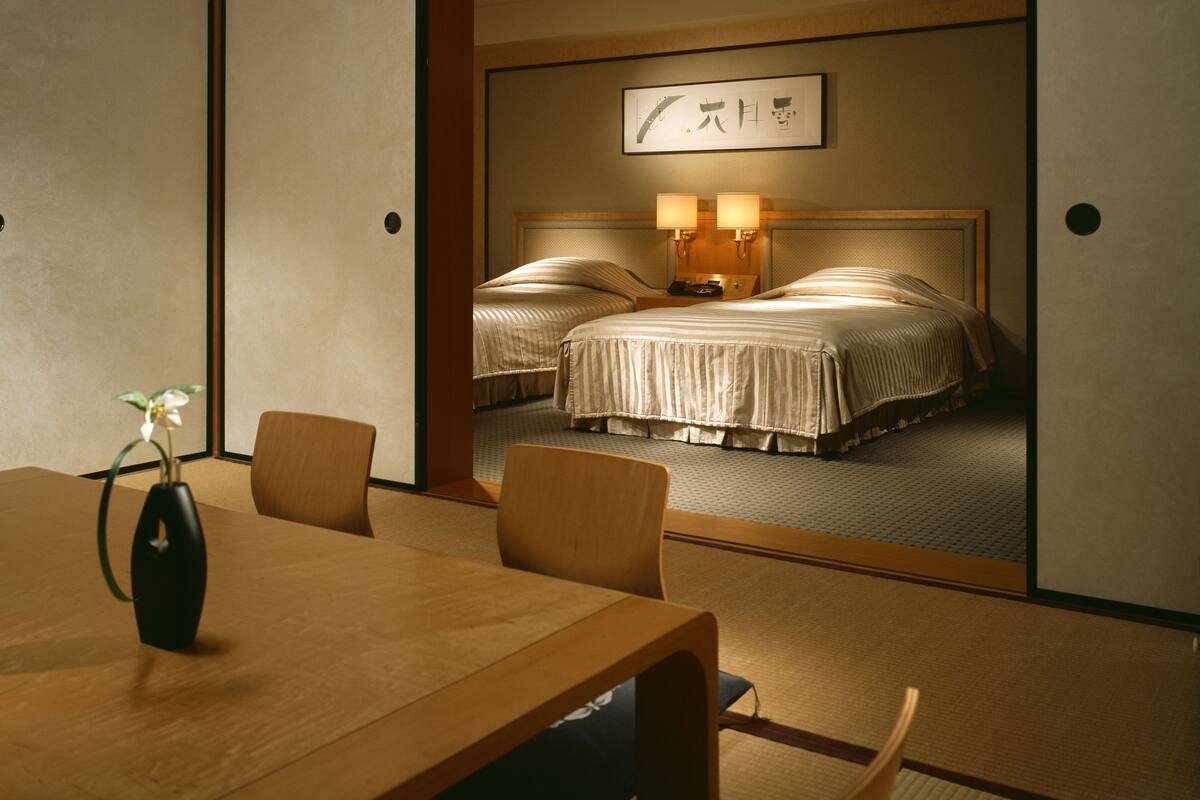 A hotel room with a traditional Japanese aesthetic, featuring tatami flooring, a low wooden table with chairs, and a bed with a headboard and bedside lamps.