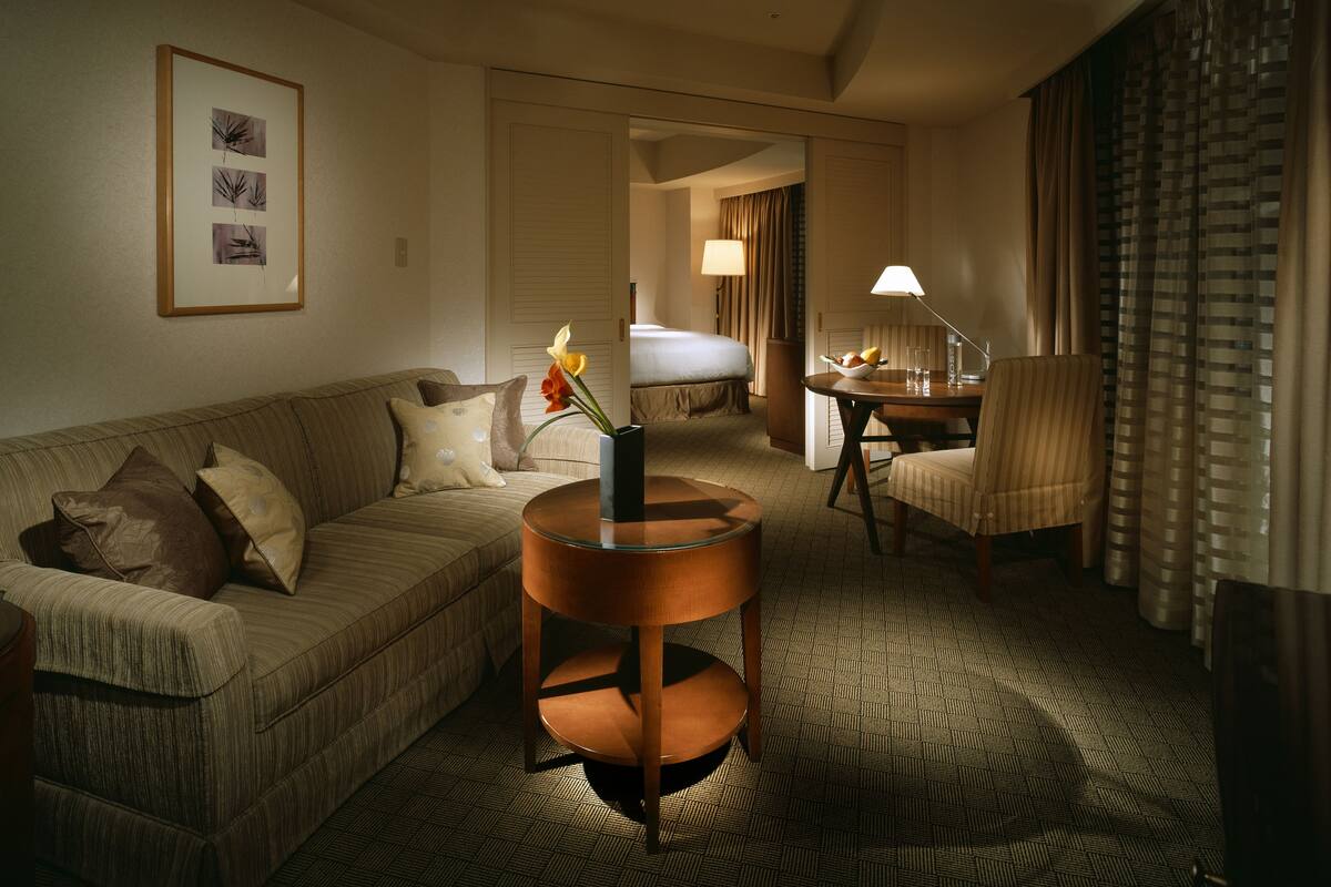 A hotel room with a sofa, a small round table with a vase of flowers, a chair, a bed, and a desk with a lamp.