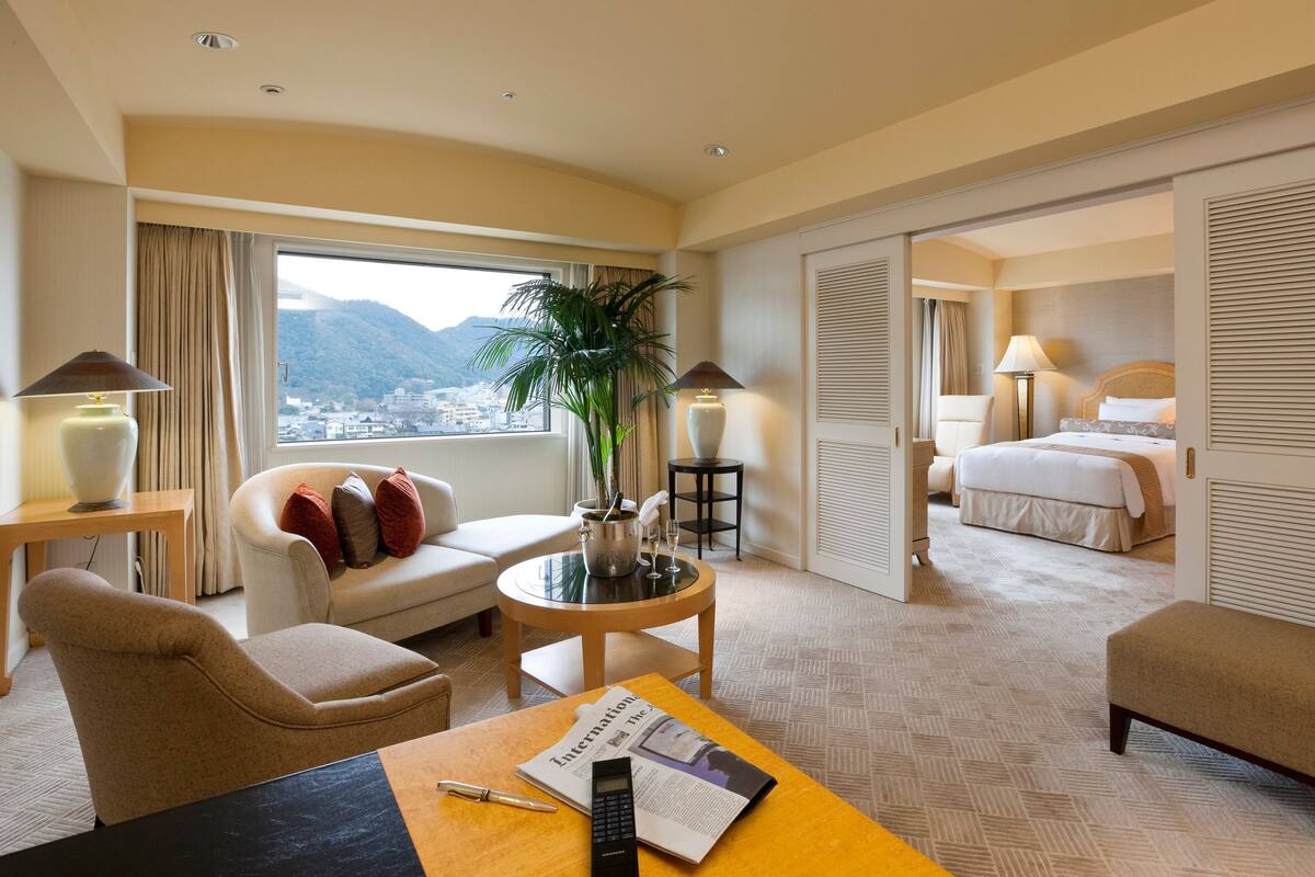 A hotel room with a sofa, armchair, and a small table with a lamp, a potted plant, and a bottle of champagne.