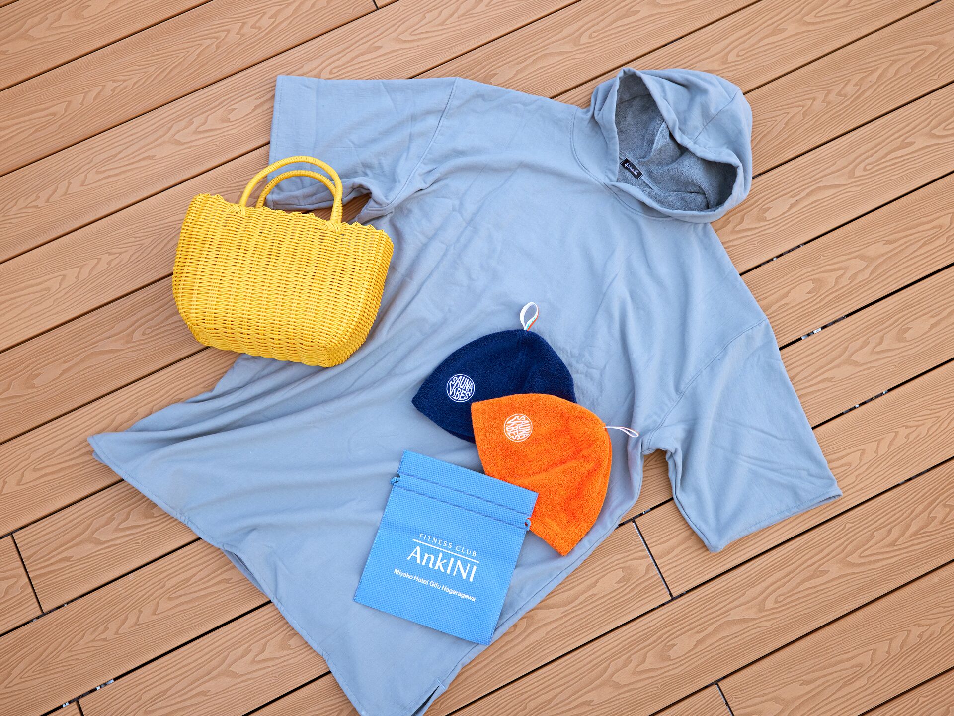 A light blue t-shirt, a yellow woven bag, two face masks, and a blue booklet on a wooden floor.
