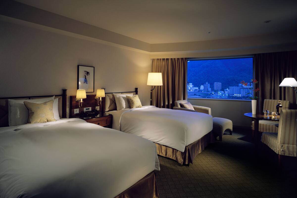 A hotel room with two beds, a desk, a chair, and a view of a cityscape at night.