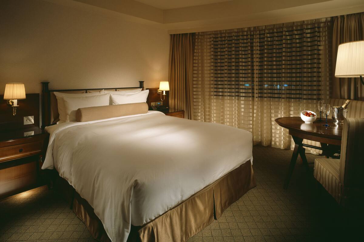 A hotel room with a large bed, two bedside tables with lamps, a small table with a bowl and glasses, and a window with curtains.