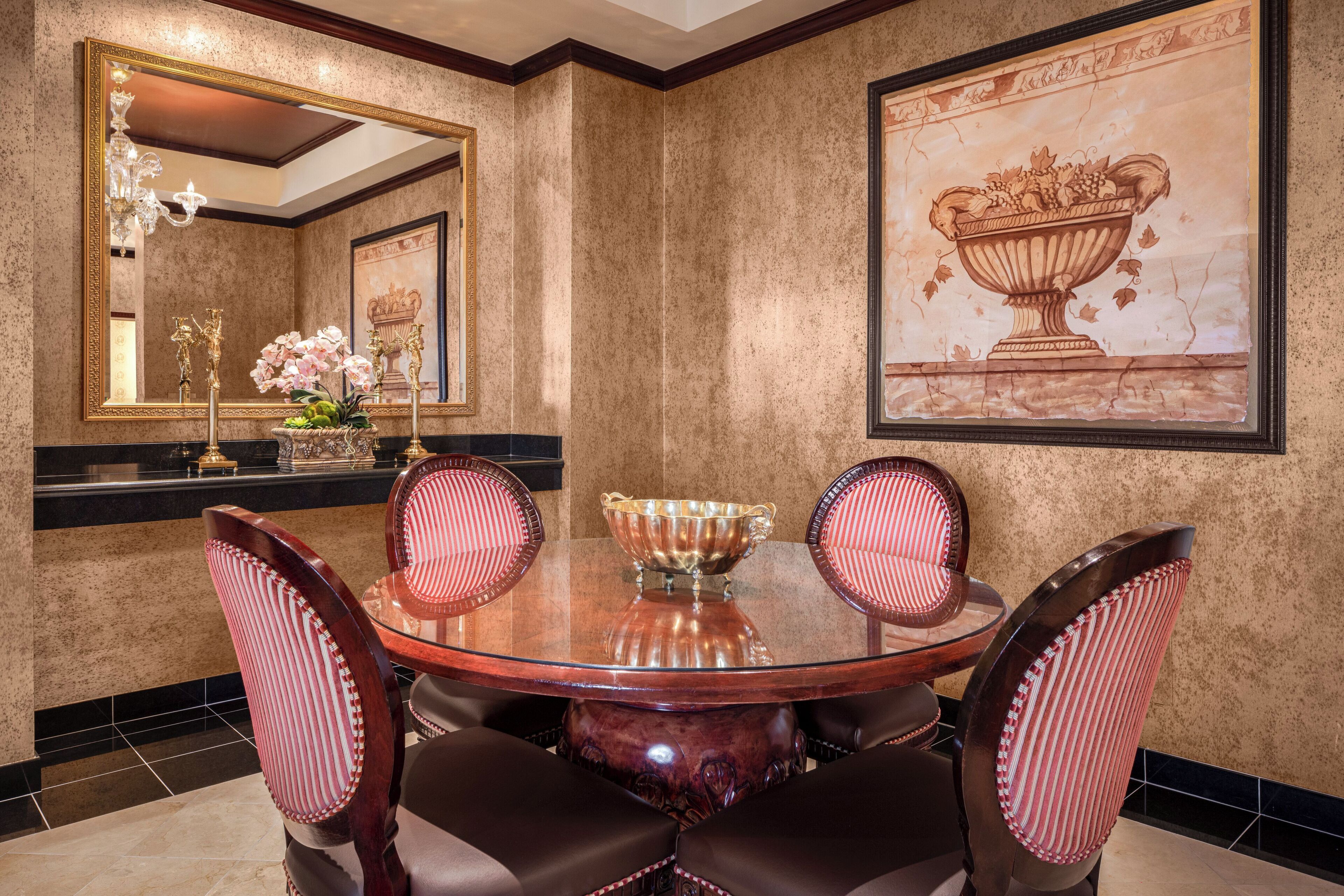 A dining area with a round glass table, four upholstered chairs, a gold bowl, and a large framed painting of a fruit bowl on the wall.