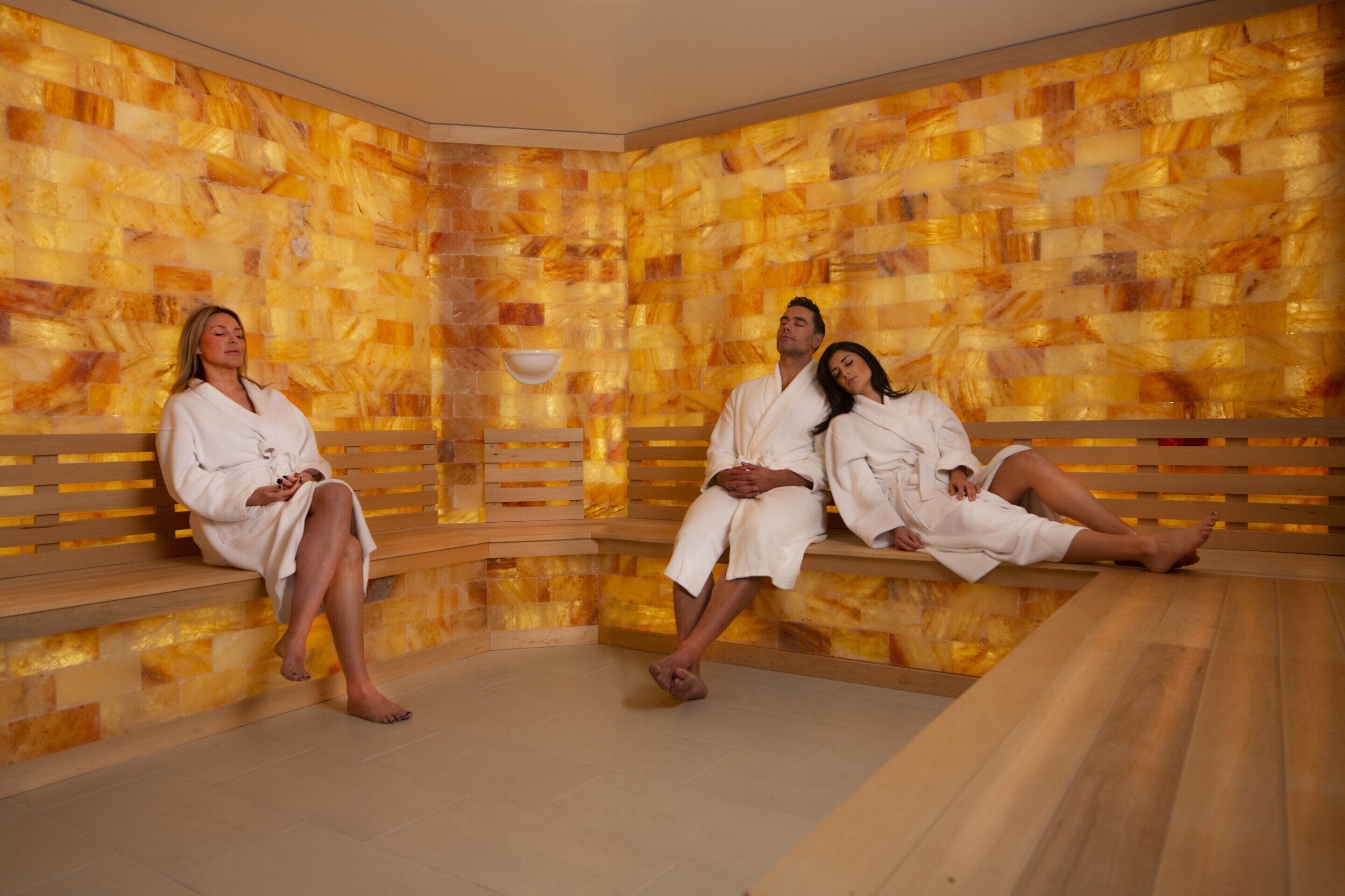 A sauna with three people in white robes, wooden benches, and a mosaic wall.