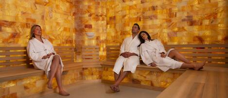 A sauna with three people in white robes, wooden benches, and a mosaic wall.