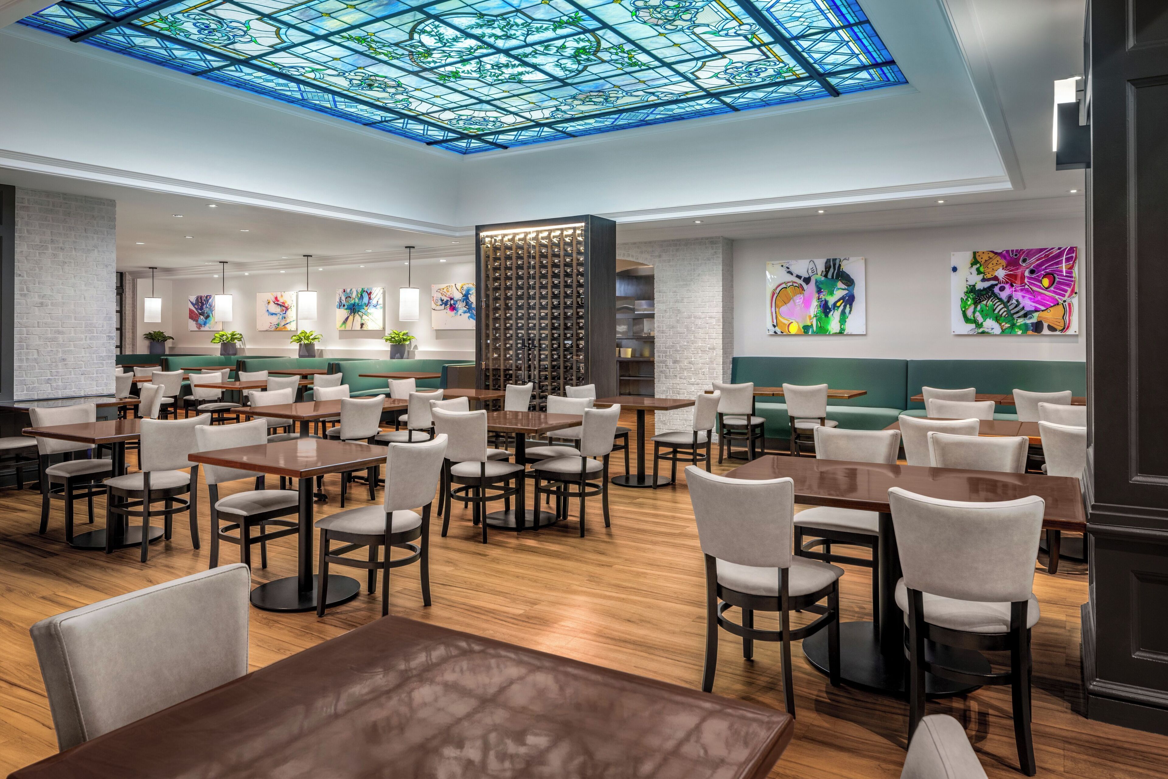 Un restaurante espacioso con numerosas mesas y sillas, un techo alto con un diseño de vitral y coloridas obras de arte en las paredes.