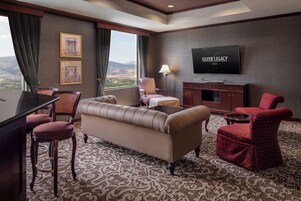 Habitación de hotel con sofá, sillones, televisor y vistas a las montañas.