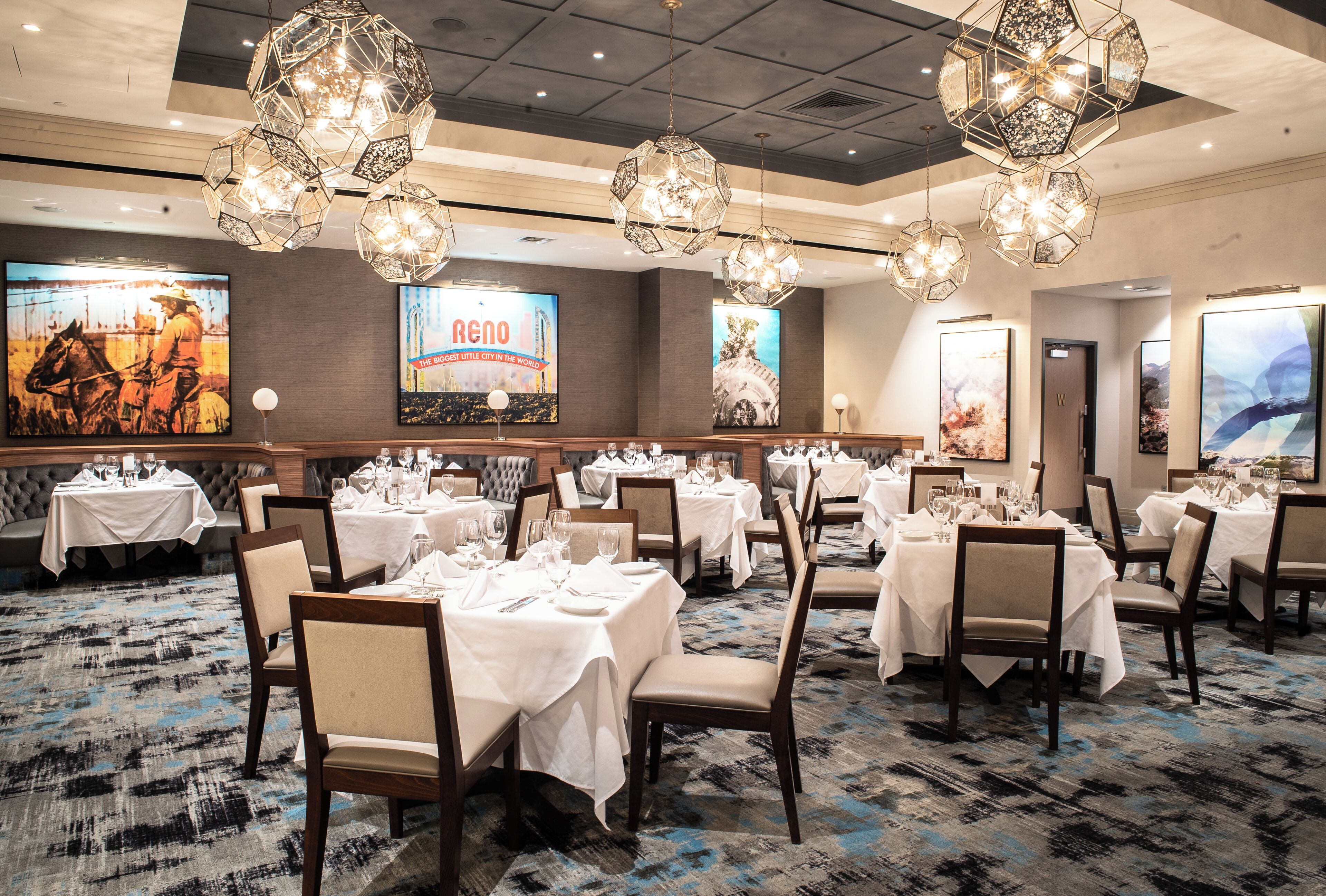 A spacious dining area with multiple tables set for dining, elegant chandeliers, and large framed artworks on the walls.