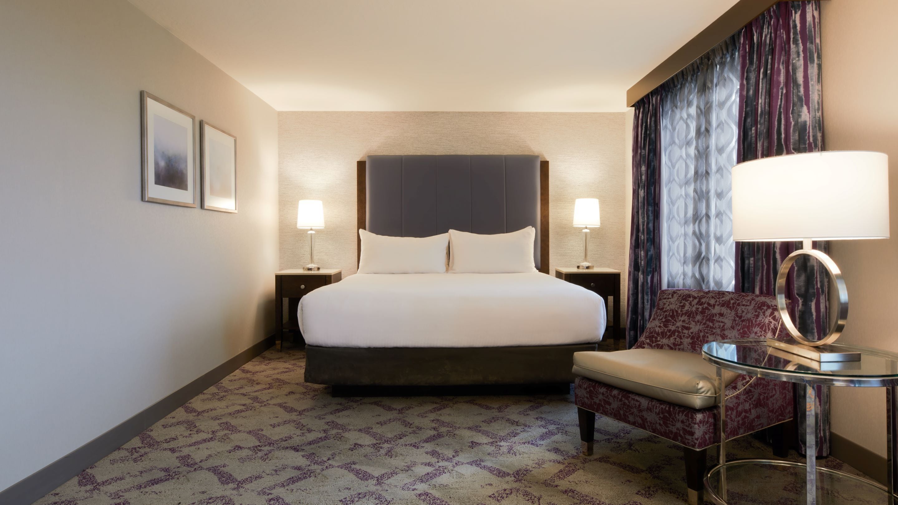 A hotel room with a large bed, two bedside tables with lamps, a chair, and a small table with a lamp.
