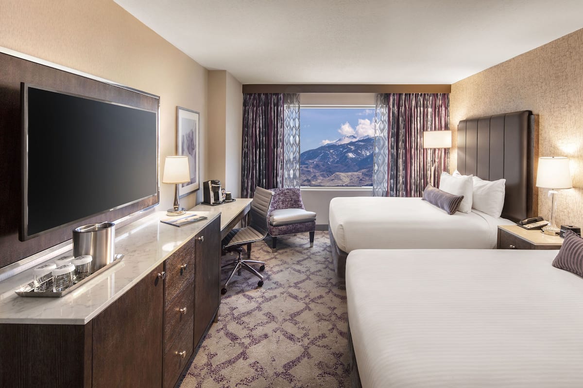 A hotel room with two beds, a desk with a flat-screen TV, a coffee maker, and a view of mountains.