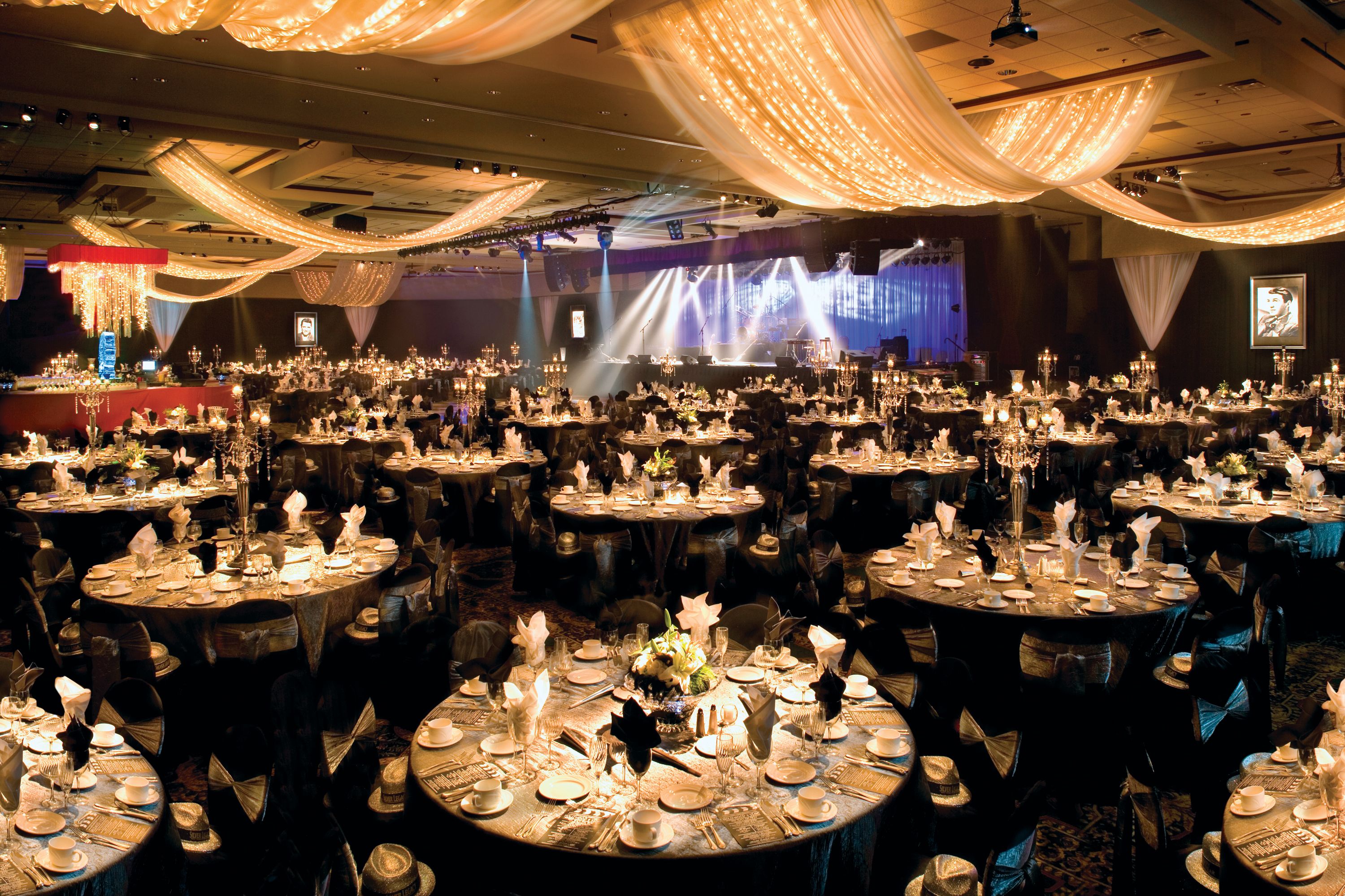 A banquet hall with round tables set for a formal event, featuring chandeliers and draped fabric.
