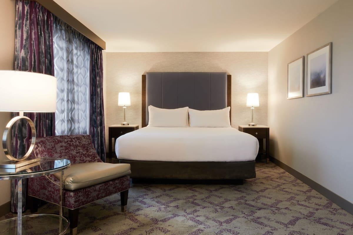 A hotel room with a large bed, two bedside lamps, a round table with a lamp, a chair, and a patterned carpet.