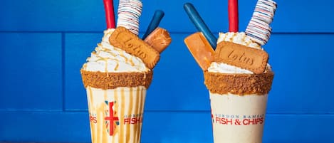 Two milkshakes with toppings and branded straws against a blue background.