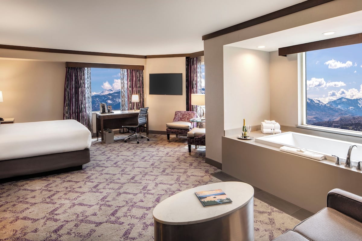 A hotel room with a bed, desk, chair, and a large window with a mountain view.
