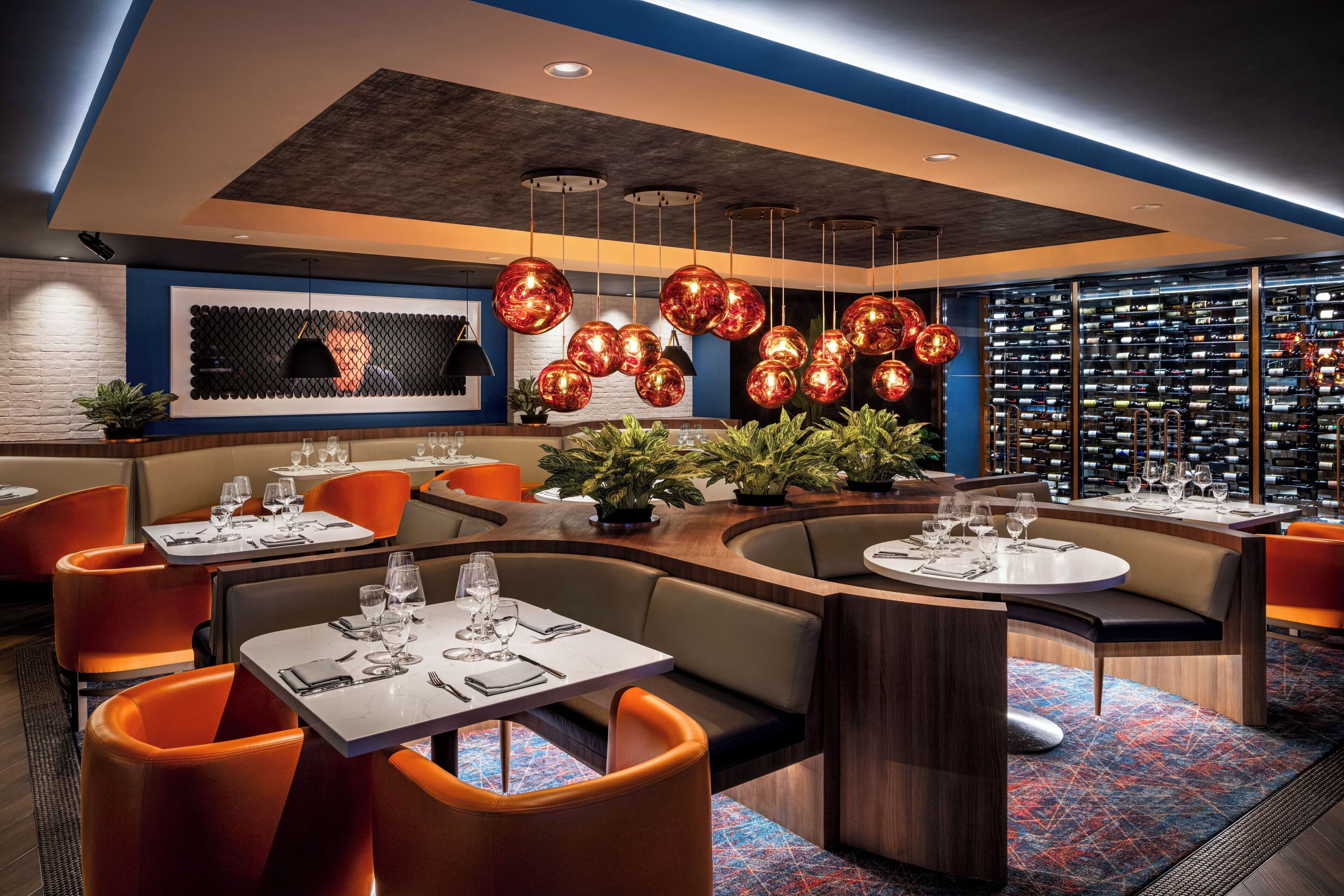 A modern restaurant with round tables, orange chairs, and a wine rack in the background.