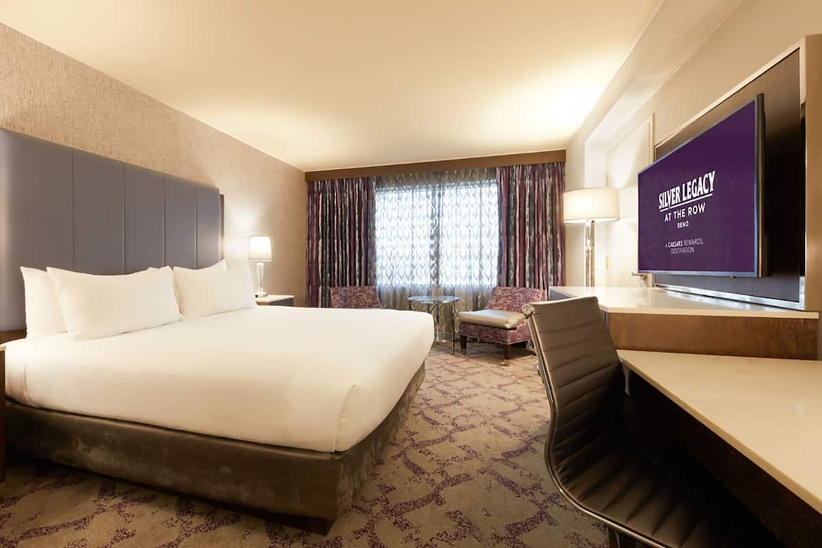 A hotel room with a large bed, two bedside tables with lamps, a desk with a chair, a television, and a patterned carpet.