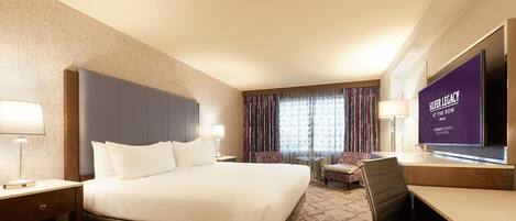 A hotel room with a large bed, two bedside tables with lamps, a desk with a chair, a television, and a patterned carpet.