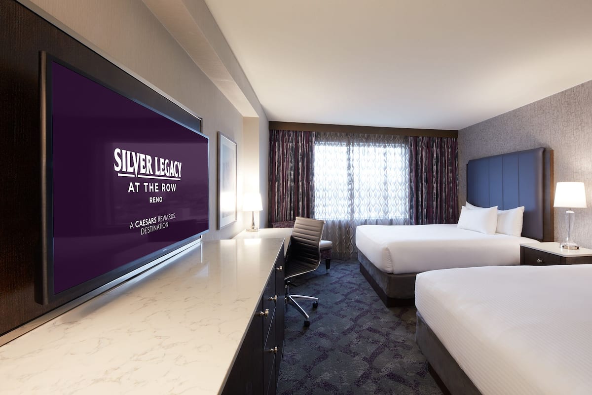 A hotel room with two beds, a desk, a chair, a TV screen displaying 'SILVER LEGACY AT THE ROW RENO', and a window with curtains.