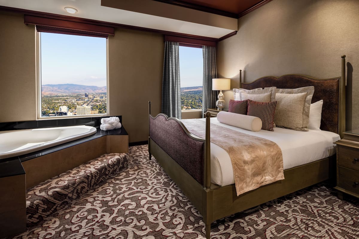 A hotel room with a large bed, a sitting area with a patterned rug, a bathtub, and a view of a cityscape through large windows.