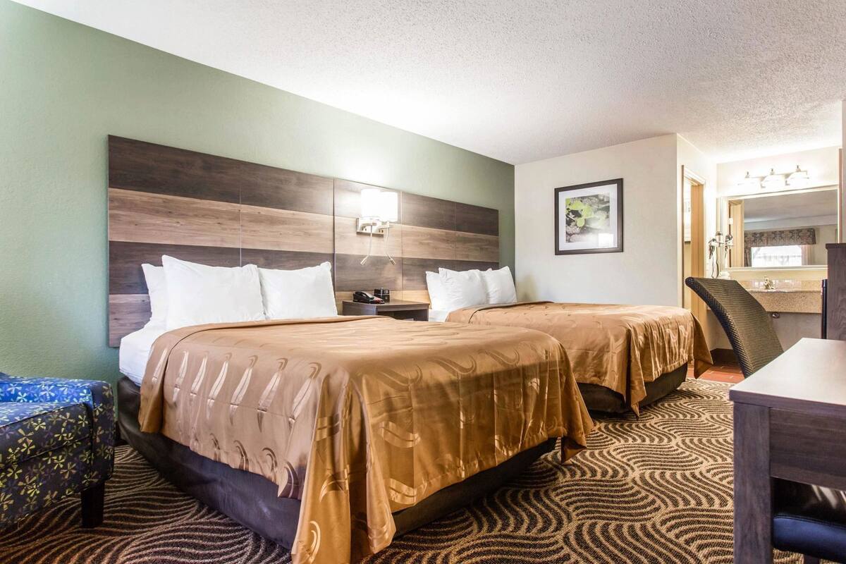 Une chambre d’hôtel avec deux lits, un bureau, une chaise, un miroir et une salle de bain visible au fond.