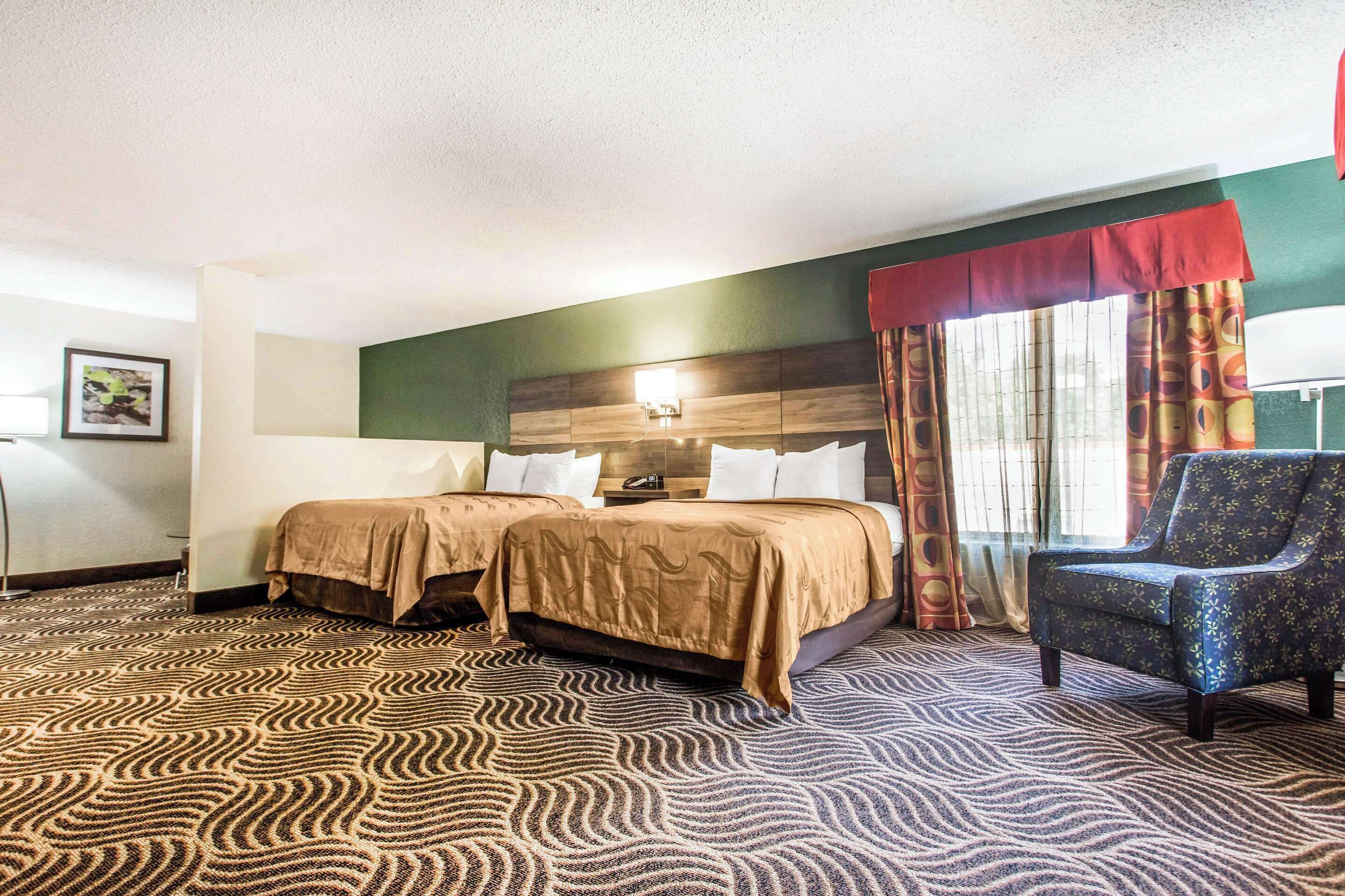 A hotel room with two beds, a chair, a patterned carpet, and a window with curtains.