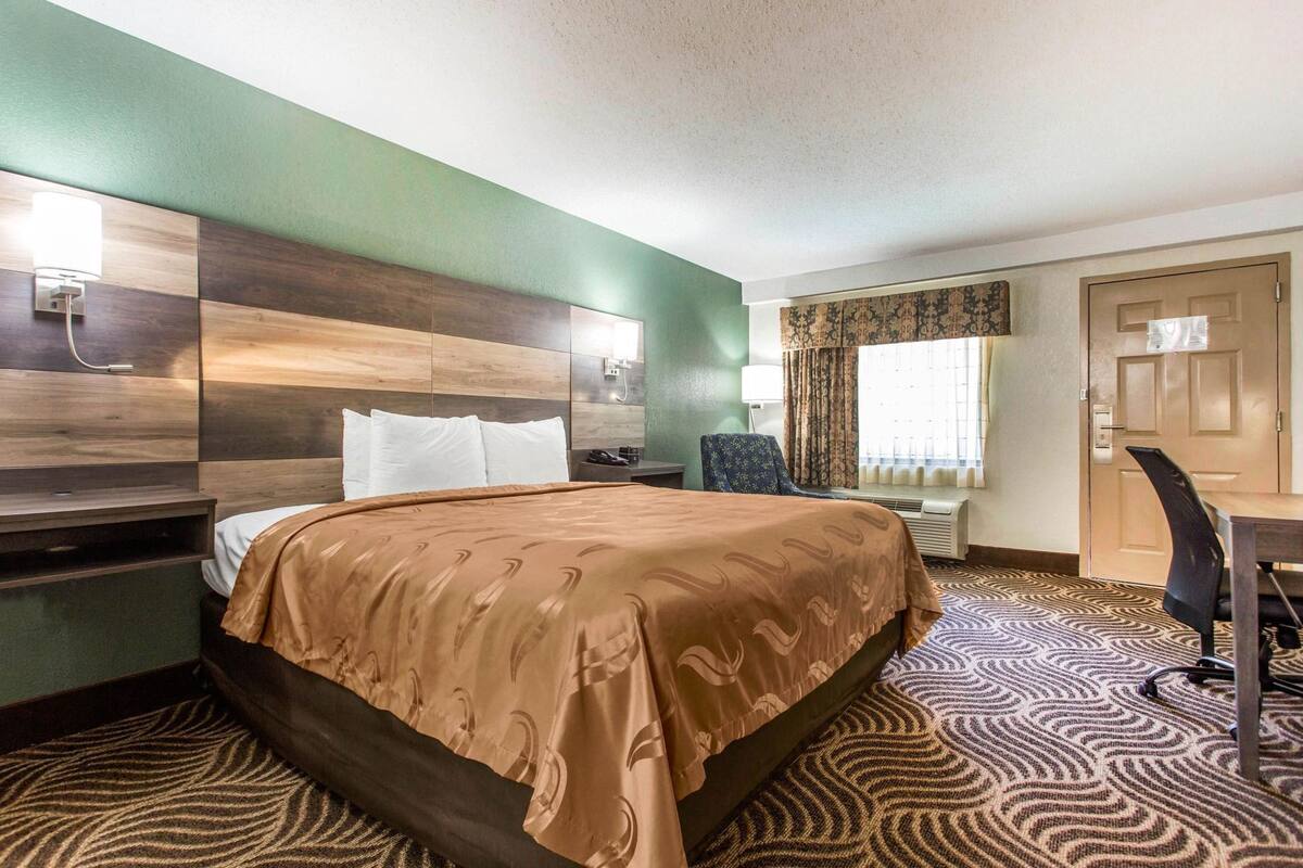 Une chambre d’hôtel avec un lit, un bureau, une chaise, une lampe et une porte.