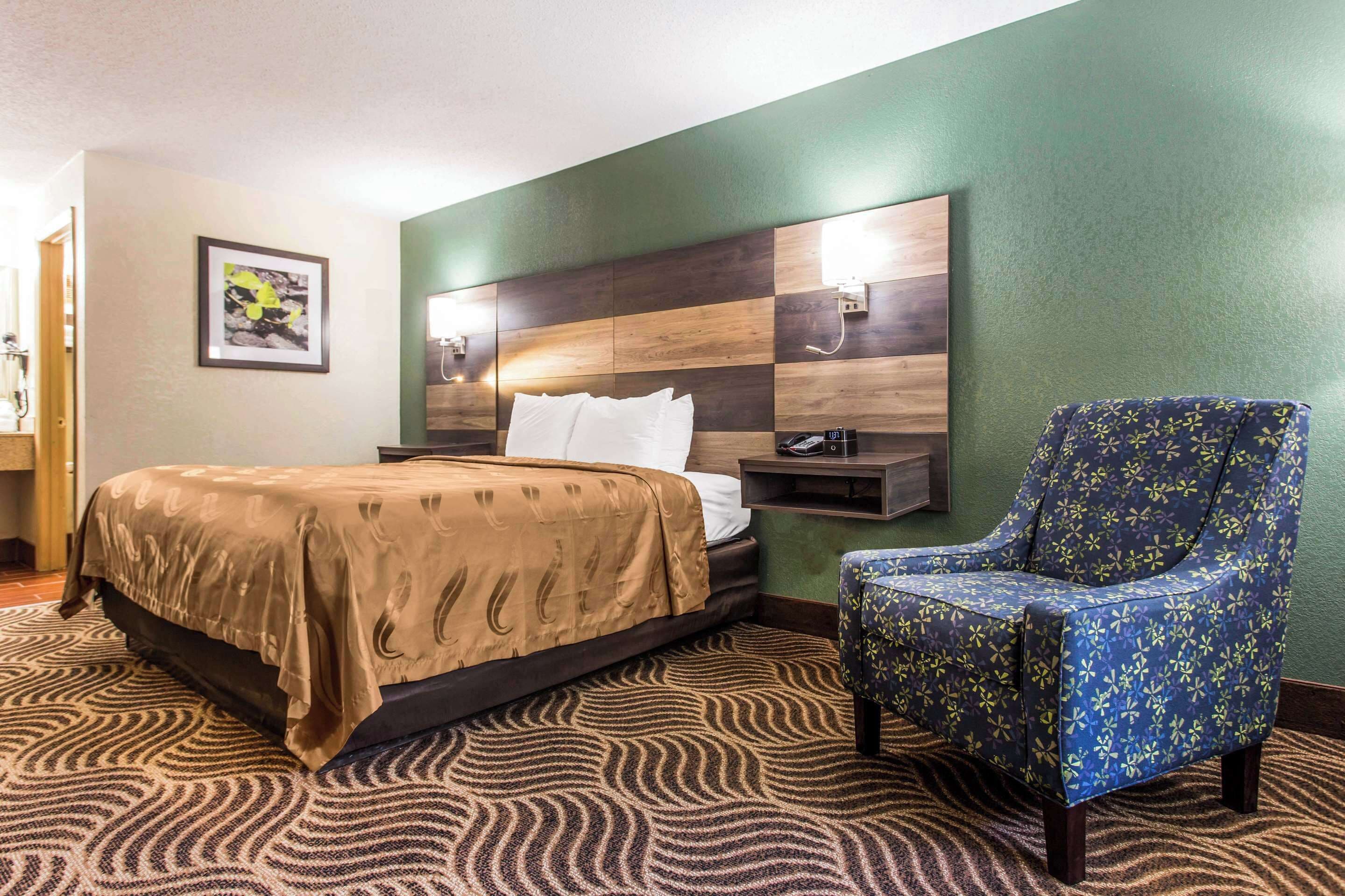 Habitación de hotel con cama, una silla, televisión montada en la pared y un cuadro enmarcado.