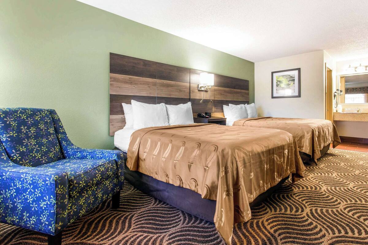 Une chambre d’hôtel avec deux lits, un fauteuil à motifs bleus, une tête de lit dotée d’un luminaire et un tableau encadré accroché au mur.