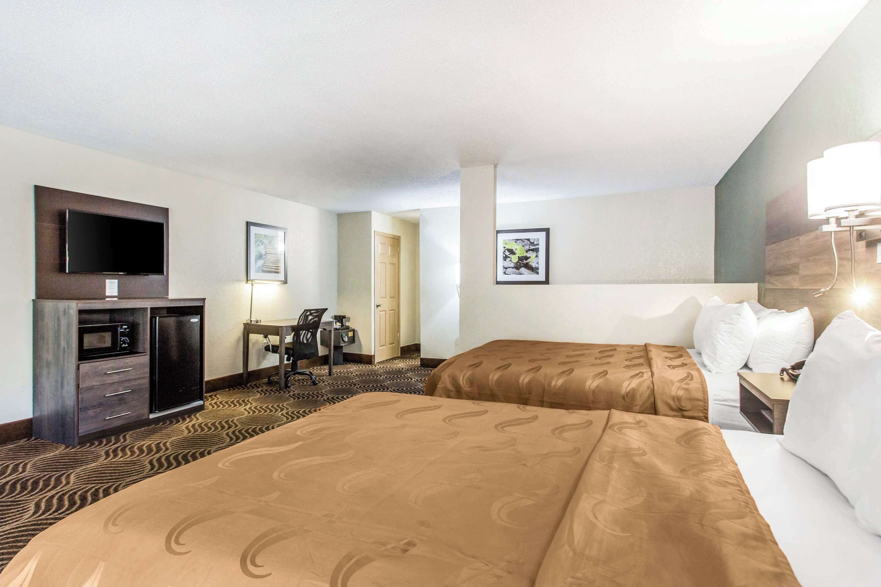 A hotel room with two beds, a desk, a flat-screen TV, a minibar, and a door leading to another room.
