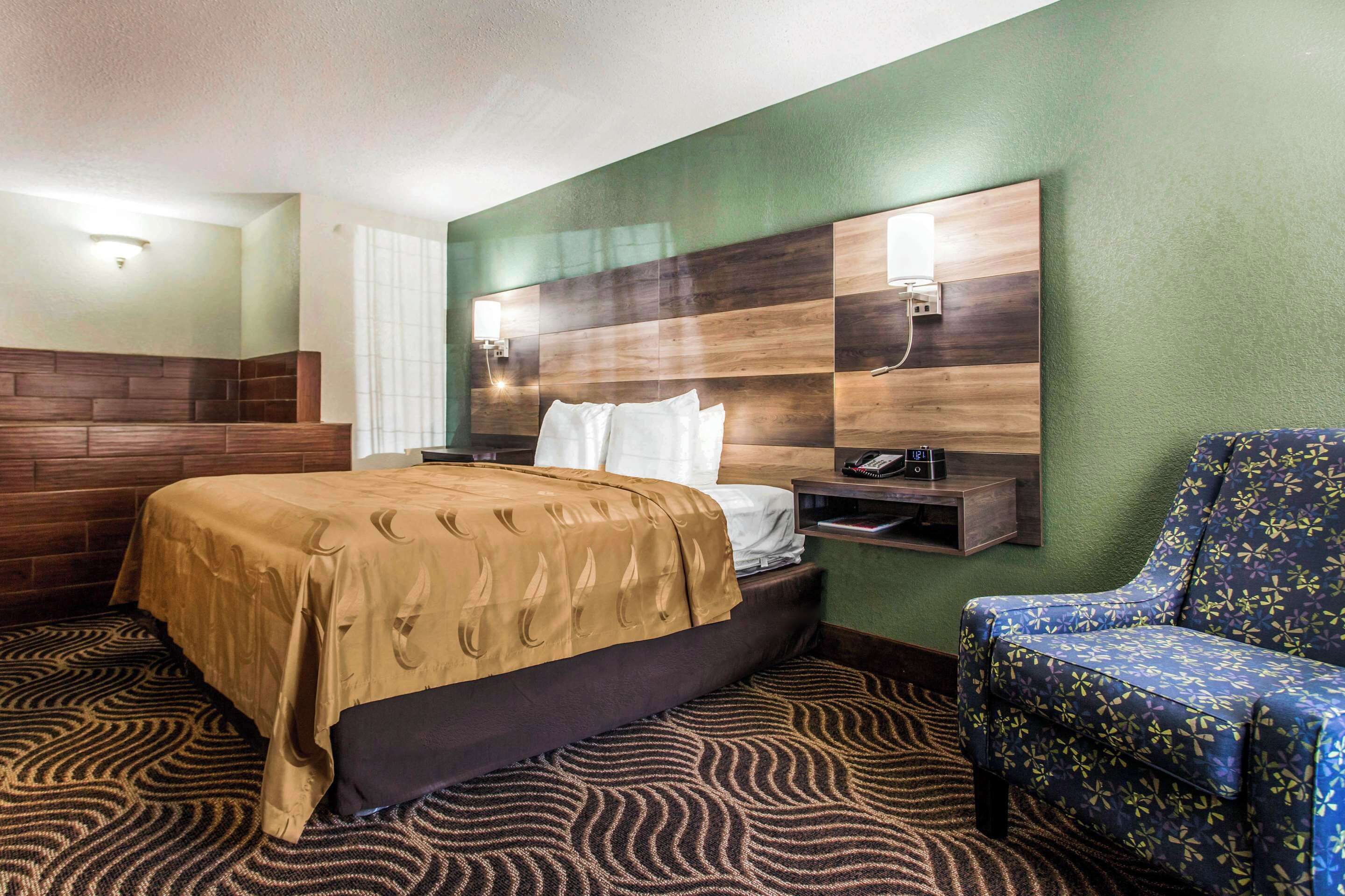 A hotel room with a bed, a chair, a nightstand, and a wall-mounted lamp.