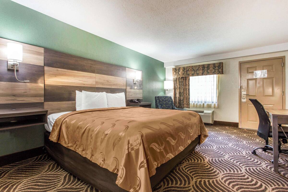 Une chambre d’hôtel avec un grand lit, un bureau avec une chaise, une table de chevet, une lampe fixée au mur et une fenêtre avec des rideaux.
