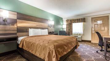 A hotel room with a large bed, a desk with a chair, a nightstand, a wall-mounted lamp, and a window with curtains.