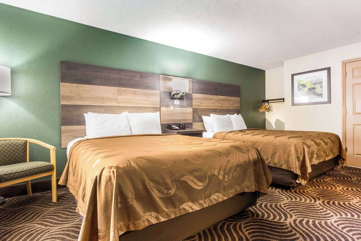 Une chambre d’hôtel avec deux lits, un mur vert, une tête de lit en bois, une chaise verte et une moquette à motifs.